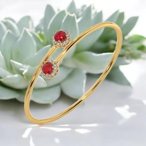 Könnte beinhalten: Ein goldfarbenes Armreif-Armband mit zwei roten Edelsteinen, die von kleinen, klaren Steinen umgeben sind. Das Armband hat ein zartes, offenes Design. Der Hintergrund zeigt eine weiche, hellgrüne Pflanze.