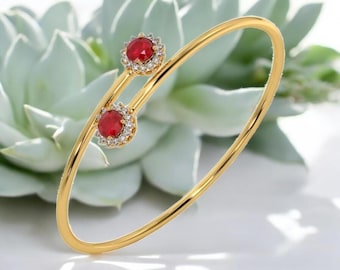 May include: A gold-colored bangle bracelet with two red gemstones encircled by small, clear stones. The bracelet has a delicate, open design. The background features a soft, light green plant.