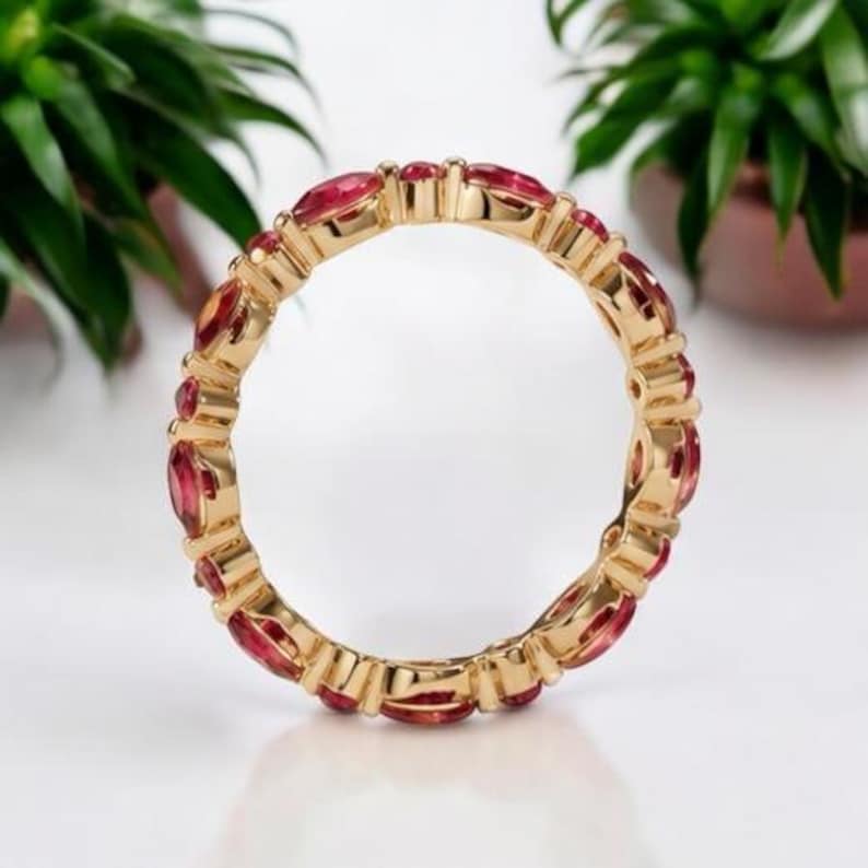 May include: A gold eternity band featuring a continuous row of marquise-cut red gemstones set in a polished yellow gold metal frame. The ring displays a repeating pattern of faceted red stones secured by gold prongs, creating a circular design.