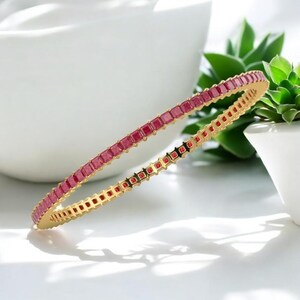 May include: A gold bangle featuring a continuous line of square, ruby-red gemstones. The bracelet is displayed against a white backdrop, with a small green plant in a white pot.