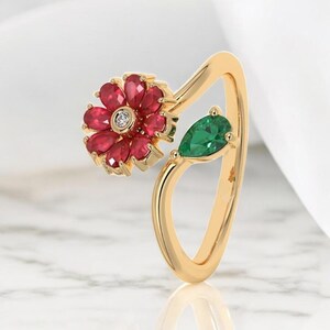 May include: A gold ring featuring a flower design with red gemstones and a central diamond. A green, pear-shaped gemstone is set on the side. The ring has a unique, open design.