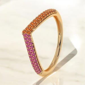 May include: A gold ring with a distinctive angular shape. The ring is embellished with small, colourful gemstones. The gemstones transition from orange to pink, creating a gradient effect. The ring is set against a white background.