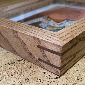 Puede incluir: Un marco de fotos de madera con un frente de vidrio y un acabado de madera marrón claro. El marco tiene una esquina biselada con un patrón de veta de madera visible.