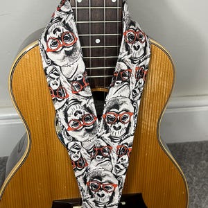 May include: A fabric ukulele strap featuring a repeating black and white illustration of monkeys wearing bright orange glasses, draped over the neck and body of a light brown wooden ukulele with a dark fretboard and silver frets.