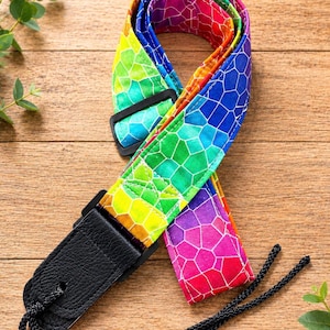 May include: A guitar strap featuring a vibrant rainbow-colored mosaic pattern with white grout lines. The strap includes black plastic adjustment buckles, black leather end tabs, and a black braided cord attachment for securing to the instrument.