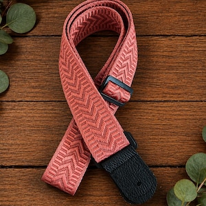 May include: A coral-colored guitar strap featuring a woven chevron pattern. The strap includes a black plastic adjustable slider and a black textured leather end tab with a single circular hole for attachment to a guitar strap button.
