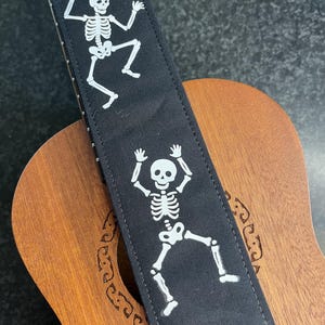 May include: A black guitar strap featuring a repeating white graphic print of dancing skeletons. The strap is positioned over the wooden body and neck of a stringed instrument, showcasing a stitched fabric construction with a clear, high-contrast design.