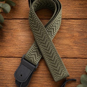 May include: An olive green guitar strap featuring a woven chevron pattern throughout the fabric. The strap includes black leather end tabs with a reinforced hole for attachment and a black plastic adjustment buckle. A thin black cord is attached.