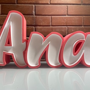 Puede incluir: Letras de espuma rosa que deletrean el nombre "Ana" en estilo cursivo. Las letras están delineadas en blanco y tienen un efecto 3D.