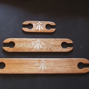 Pode incluir: Três lançadeiras de tecelagem de madeira com um design branco em cada uma. As lançadeiras são de tamanhos diferentes, sendo a maior na parte inferior e a menor na parte superior.