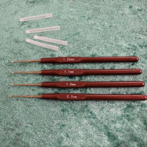 May include: Four crochet hooks with dark red handles and silver tips, labeled 1.25mm, 1.1mm, 1.0mm, and 0.9mm. Several small, clear plastic thread protectors are also present. The tools are for crafting.
