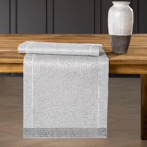 Könnte beinhalten: Ein weißer und grauer Tischläufer mit einem dekorativen Rand. Der Läufer ist in der Mitte gefaltet und auf einem Holztisch platziert.