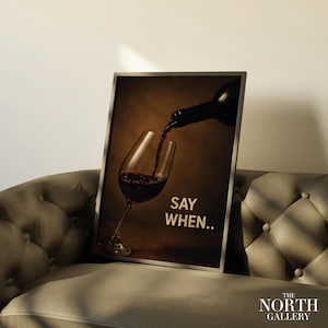 May include: A framed print featuring a wine glass being filled with red wine from a dark bottle. The words "SAY WHEN.." are printed in white. The North Gallery logo is in the bottom right corner. The print is displayed on a tufted sofa.