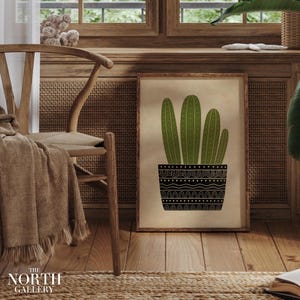 May include: A framed art print depicting a green cactus in a black patterned pot. The artwork is displayed in a room with wooden furniture and flooring. The print is from The North Gallery.