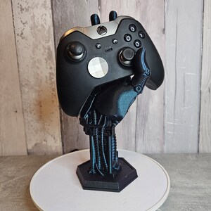 May include: A black and silver video game controller is held by a 3D printed robotic hand, which is mounted on a white circular base.
