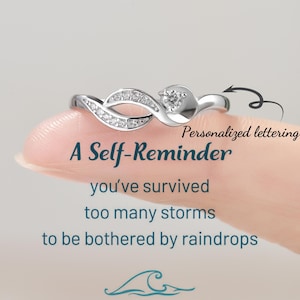 May include: A silver ring with a small diamond and a wave design. The ring has the inscription "A Self-Reminder you've survived too many storms to be bothered by raindrops".