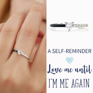 May include: A silver ring with a heart-shaped diamond and small accent stones. The ring has a black band on one side. Text on the right says "A SELF-REMINDER" and "Love me until I'M ME AGAIN".