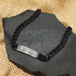 May include: A black chain bracelet with a rectangular tag featuring a black and white photo of a person's eyes.