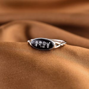 May include: A silver ring with a black oval centerpiece featuring white floral designs. The ring has a delicate band and is set against a brown fabric background. The floral designs are detailed and elegant.