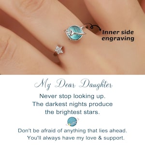 May include: A silver ring set with a blue planet design and a star. The ring has the words "My Dear Daughter" and a message about looking up and love. The ring is shown on a finger.