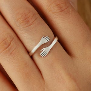 May include: A silver ring with two hands forming a hug. The ring is open and adjustable to fit different finger sizes.