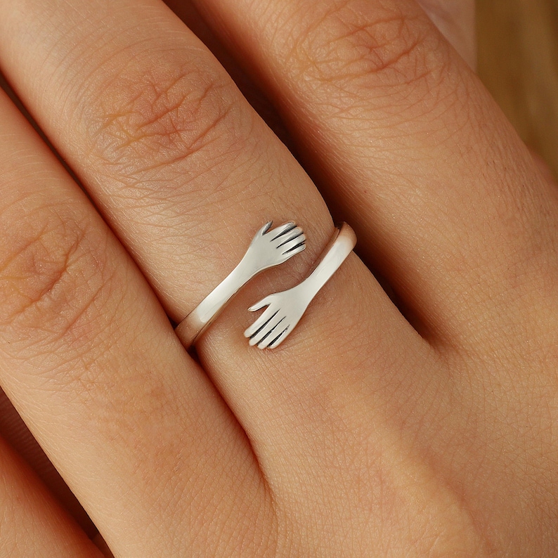 May include: A silver ring with two hands forming a hug. The ring is open and adjustable to fit different finger sizes.