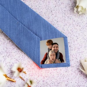 May include: A light blue tie with a textured pattern. The tie features a rectangular photo of three people. The photo shows a man with a child on his shoulders and an older man in front.
