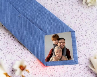 May include: A light blue tie with a textured pattern. The tie features a rectangular photo of three people. The photo shows a man with a child on his shoulders and an older man in front.