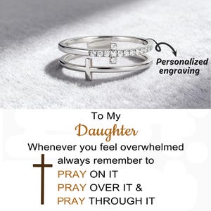 Peut inclure: Une bague en argent avec un motif de croix. La bague a deux bandes, l'une ornée de petites pierres claires. L'image comprend le texte "To My Daughter" et "Gravure personnalisée". C'est un bijou à thème religieux.