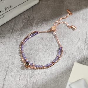 May include: A rose gold bracelet with two strands. One strand features small, round beads, and the other has small, faceted purple beads. The bracelet has a sliding clasp for adjustable sizing.