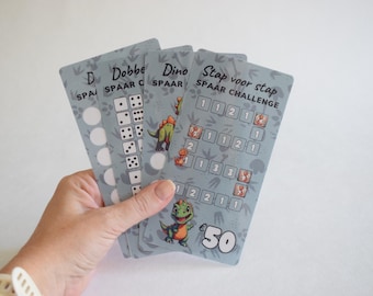 May include: Four cards with a dinosaur theme. The cards are for a savings challenge. The cards have different tasks to complete, such as rolling dice, saving money, and completing a step-by-step challenge. The cards are blue and grey with illustrations of dinosaurs and dice.