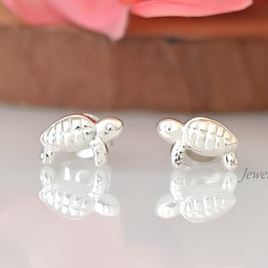 Könnte beinhalten: Ein Paar silberne Ohrstecker in Form von Schildkröten. Jeder Ohrring hat ein detailliertes Schildkröten-Design mit strukturiertem Panzer und kleinen Beinen. Die Ohrringe werden auf einer reflektierenden Oberfläche mit rosa Blumen im Hintergrund präsentiert.