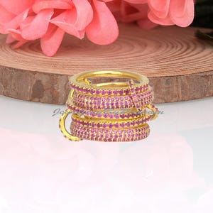 Op de afbeelding: Een goudkleurige ring met meerdere gestapelde banden bezet met kleine, levendige roze steentjes. De ring staat voor een bos koraalroze bloemen en rust op een ronde, lichtbruine houten schijf als ondergrond.
