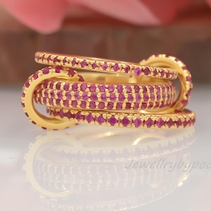 May include: A gold-colored ring set with multiple bands, each encrusted with small, vibrant red gemstones. The ring is displayed against a blurred background of pink flowers, creating a striking contrast. The ring's design is intricate and eye-catching.