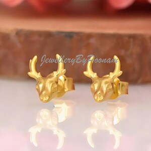 May include: A pair of gold-colored stud earrings shaped like deer heads. The earrings are displayed on a reflective surface, with a blurred background of pink flowers and a wooden disc. The earrings are small and detailed.