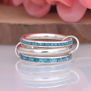 May include: A silver ring set consisting of three interlocking bands. Two of the bands feature a channel setting with small, square-cut, light blue gemstones, while the central band is a plain, polished silver ring without any stones.