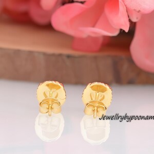 May include: Close-up of two gold-colored stud earrings with a round, textured design. The earrings have a post back and are set against a blurred background with pink flowers and a wooden surface. The text "Jewellrybypoonam" is visible.