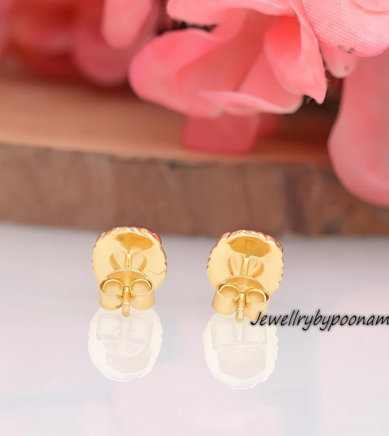 May include: Close-up of two gold-colored stud earrings with a round, textured design. The earrings have a post back and are set against a blurred background with pink flowers and a wooden surface. The text "Jewellrybypoonam" is visible.