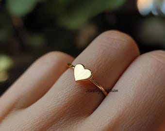 May include: A gold-colored ring featuring a heart-shaped design. The ring is simple and elegant, with a thin band and a flat heart. The text "Jewellrybypopham" is visible on the ring.