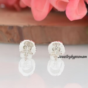 May include: A pair of silver stud earrings with a round, textured design and a red center. The earrings have a post and butterfly backing. The text "Jewellrybypoonam" is visible on the right side of the image.