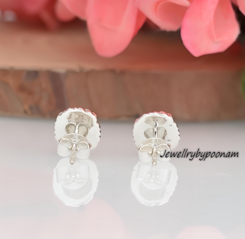 May include: A pair of silver stud earrings with a round, textured design and a red center. The earrings have a post and butterfly backing. The text "Jewellrybypoonam" is visible on the right side of the image.