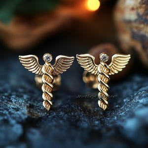 Peut inclure: Une paire de boucles d'oreilles en or avec un motif de caducée ailé et un petit strass transparent au sommet de chaque boucle d'oreille.