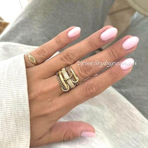 Op de afbeelding: Een hand met een gouden ring met een klein ovaal ontwerp en een gelaagde gouden ring met zwarte en heldere stenen. De nagels zijn lichtroze geverfd. De tekst "Jewellrybypoon.am" is zichtbaar.
