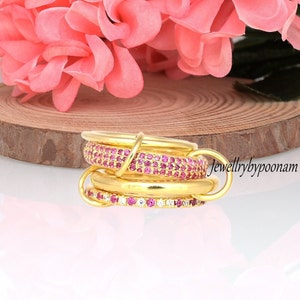 May include: A set of four gold rings with pink and white stones. The rings are stacked on top of each other and are connected by a small gold loop. The rings are on a wooden surface.  "jewellerybypoonam"