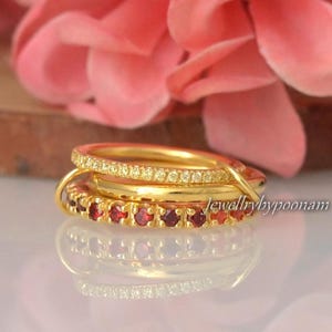 Puede incluir: Tres anillos de oro apilados. El anillo superior está incrustado con pequeñas piedras transparentes. El anillo del medio es de oro liso. El anillo inferior está adornado con pequeñas piedras rojas. Los anillos están sobre un fondo rosa borroso.