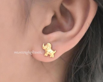 May include: A gold-toned stud earring shaped like a triceratops dinosaur. The earring features a textured surface depicting the dinosaur's three horns, frill, and tail, securely fastened to the earlobe. The metal has a polished, reflective finish.