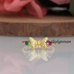 Könnte beinhalten: Goldener Schmetterlingsring mit grünen und roten Edelsteinen. Der Schmetterling hat klare Steine auf seinen Flügeln. Der Ring liegt auf einer Holzoberfläche. "Jewellerybypoonam" ist auf der Oberfläche geschrieben.