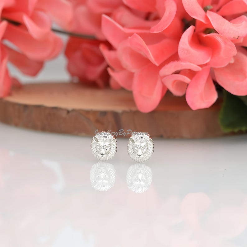 May include: A pair of silver-colored lion head stud earrings. The earrings are round with detailed lion faces. The background features pink flowers and a wooden surface.