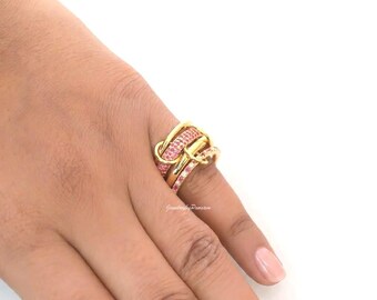 Può includere: Un anello in oro dal design intrecciato a più livelli. La fascia è composta da diversi anelli sovrapposti, con una sezione incastonata di piccole pietre rosa brillante che conferiscono un aspetto strutturato e scintillante al metallo dorato lucido.