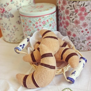 Könnte beinhalten: Nahaufnahme eines Weidenkorbs mit Filz-Croissants mit braunen Streifen. Dekorative Dosen mit Blumenmuster im Hintergrund. Die Szene spielt sich auf einer weißen Tischdecke ab, mit Keramikgriffen im Vordergrund.